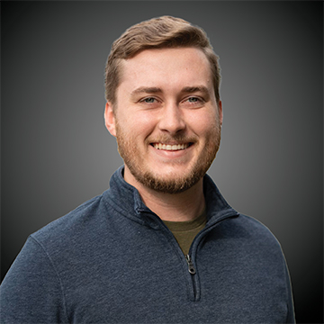 Jimmy Hartley, a man with light brown hair and a beard, smiles at the camera in a dark zip-up sweater over a green shirt, set against a plain dark gradient background.