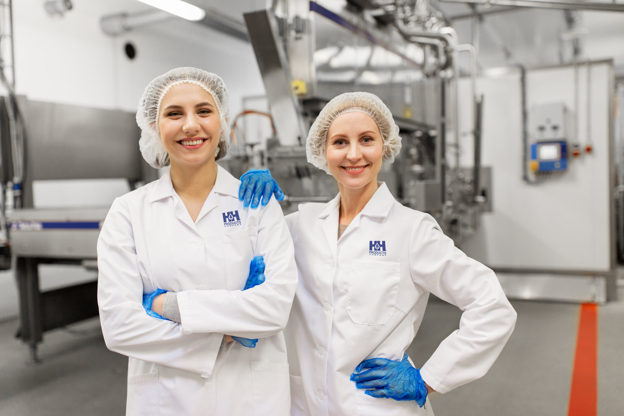 Two women wearing hair nets, white lab coats, and blue gloves stand in a food processing facility, smiling at the camera with machinery in the background.