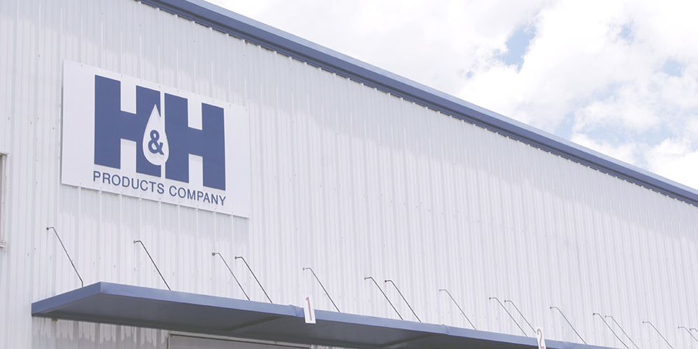 Exterior of a white industrial building with a blue H&H Products Company sign on its corrugated wall under a partly cloudy sky.
