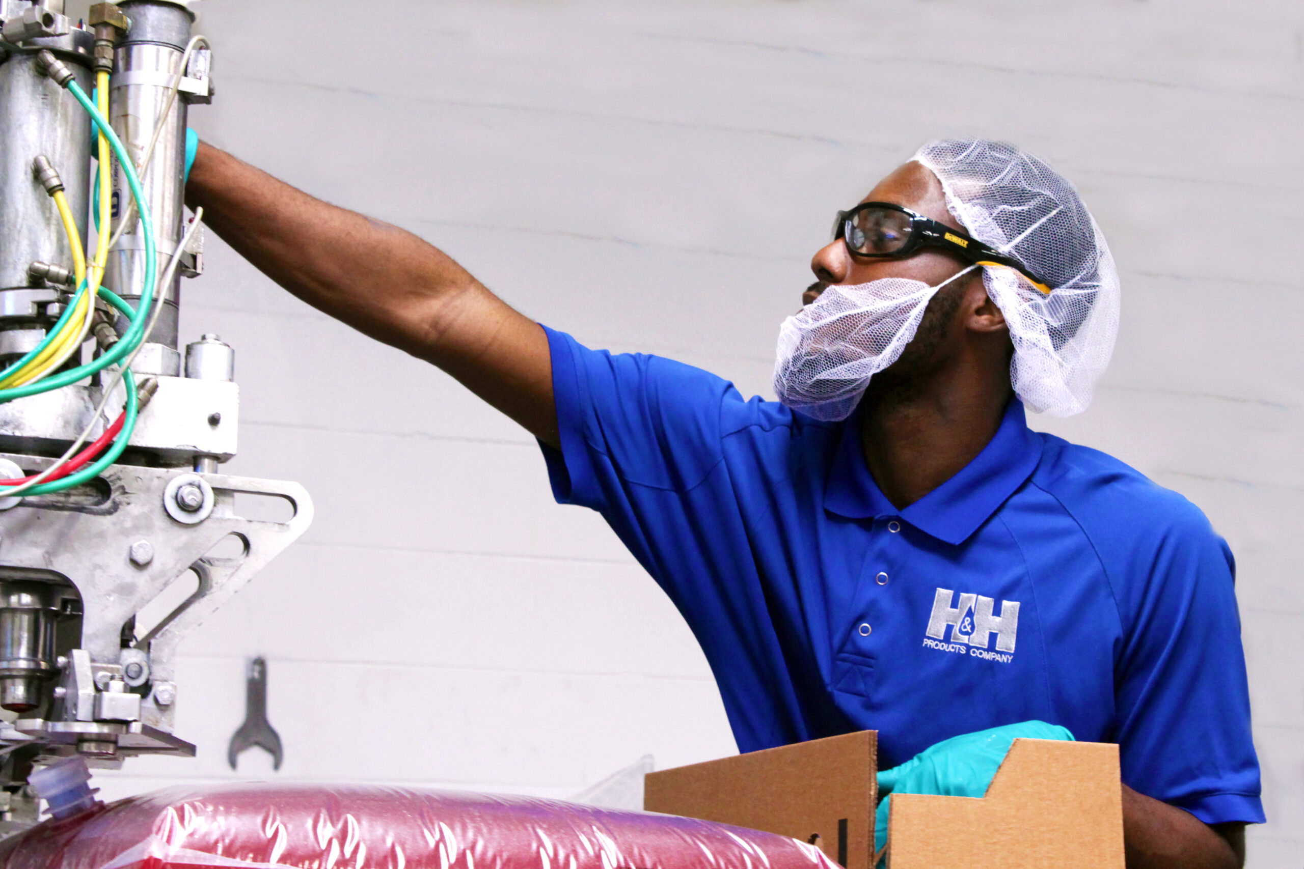 A worker wearing safety glasses, a hairnet, and gloves operates machinery in a factory setting while holding a cardboard box.