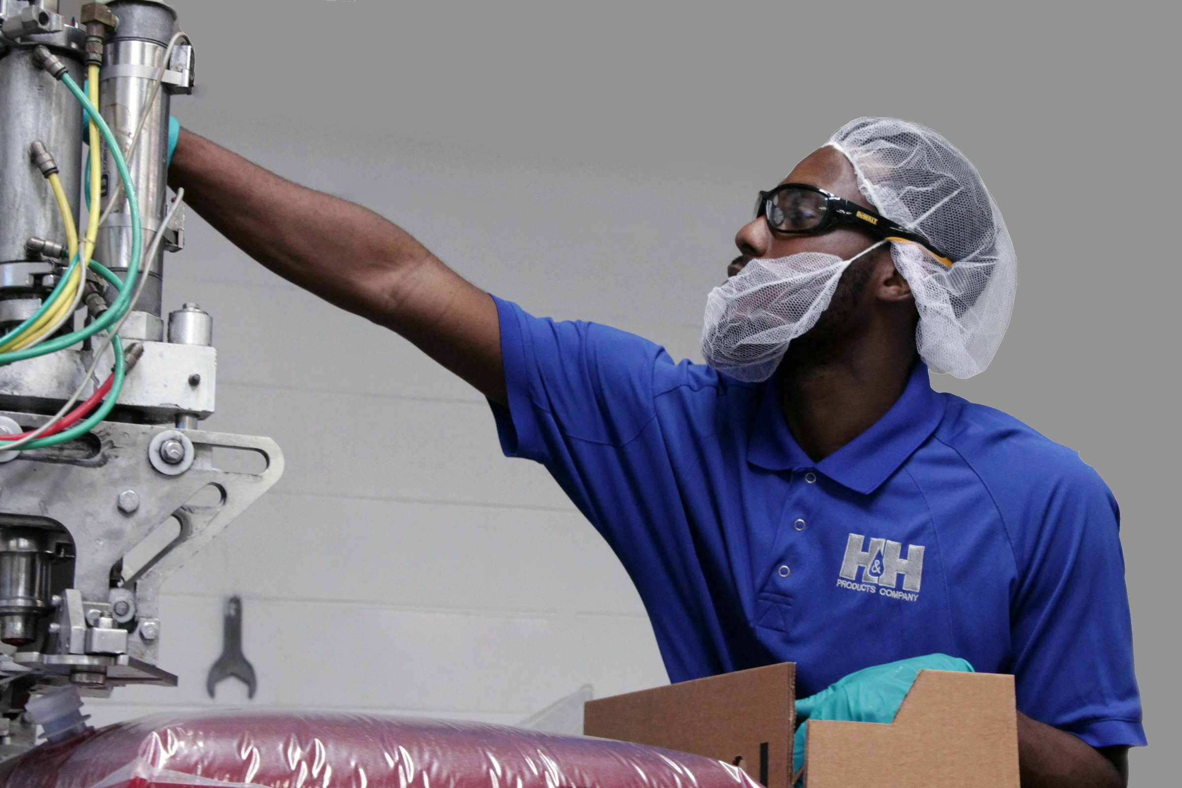 A man wearing safety glasses, a hairnet, and gloves operates industrial equipment while packing items in a factory setting.